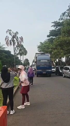 A video shows a woman wearing a pink and white hooded jacket, a maroon skirt, and a white hat standing on a paved path. She is facing to her left, with her mouth slightly open. To her left, another woman in a black hijab and a striped shirt is holding a phone, appearing to take a photo or video. In the background, a large blue truck with "PRIORITAS" written on the side is driving on the road, along with other vehicles. Palm trees and lush greenery are visible on the left side of the frame. The sky is overcast. The scene appears to be outdoors in daylight.