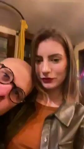 A woman with long brown hair, wearing a brown top and a green leather jacket, is filming a selfie video with a bald man with glasses. The man rests his head on her shoulder and looks at the camera. The woman laughs and says "Tu es l'handicapé" or "You are the disabled one." The background shows a window frame and a yellow curtain.