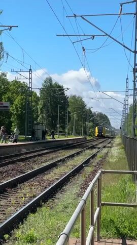 The video shows a train station platform on a sunny day. Several people are standing on the platform, waiting. One person is holding a bicycle. A yellow train is approaching the station on the tracks. Overhead are numerous power lines and supporting structures for the railway. Grass grows along the tracks and a guardrail is visible in the foreground. Trees are visible in the background.