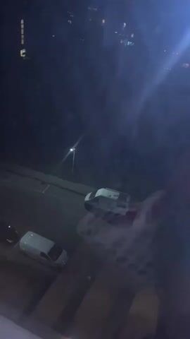 The video begins with a nighttime view from an upper story, overlooking a street with a few vehicles and streetlights. The scene then shifts to an indoor setting, possibly a hallway or laundry area. A person is seen walking while carrying a white laundry basket filled with clothes, including red, pink, and beige items. They are wearing shorts and pink slippers. The floor appears to be tiled or linoleum. The subsequent shots show a dimly lit hallway with yellow walls and a dark-colored rug on the floor. Further down the hallway, another doorway is visible, leading to a darker space, potentially an apartment or room. The overall impression is of someone moving through a residential building at night, perhaps to do laundry. No text is visible in the media.