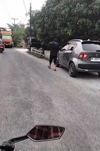 A person wearing a black shirt and pants is standing next to a car. A motorcycle is driving on a paved road. There are trees and buildings on the side of the road.