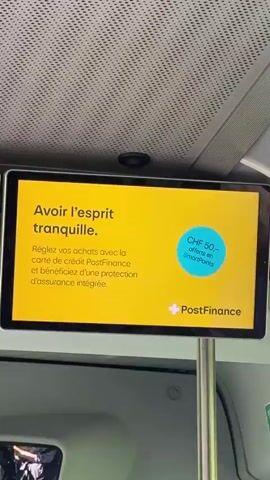 The video shows a close-up shot of a digital screen mounted inside what appears to be a public transport vehicle, possibly a bus or a train. The screen is displaying an advertisement in French. The advertisement has a bright yellow background. In the top left corner, there is text that reads "Avoir l'esprit tranquille." Below this, it says "Réglez vos achats avec la carte de crédit PostFinance et bénéficiez d'une protection d'assurance intégrée." On the right side of the screen, there is a light blue circular icon with text that appears to say "CHF 50 offert en SmartPoints." At the bottom right of the screen, there is a logo that includes the words "PostFinance". The screen is attached to a silver metal pole. The interior of the vehicle is dimly lit, with some overhead lighting visible, and parts of the ceiling and a metal support structure are in the background. There are no people visible in the frame.