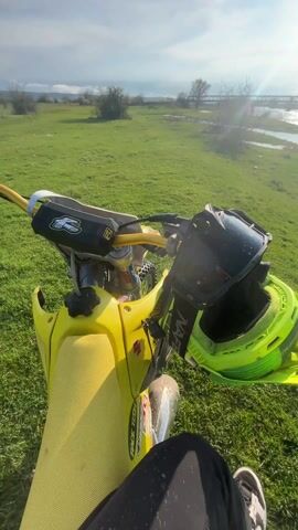 The video shows a first-person perspective from someone sitting on a yellow Suzuki dirt bike. The bike is parked in a grassy field near a body of water with a bridge in the background. The day appears sunny with some clouds. A helmet and goggles are resting on the front of the bike, obscuring the handlebars. The person is wearing dark pants and dirty sneakers. The video starts with a close-up of the helmet and then pans out to show the surrounding landscape.