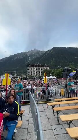 A crowd of people are gathered in an outdoor plaza, seated on wooden benches. They are watching an event, possibly a competition or performance, as indicated by the stage, sound equipment, and banners visible in the background. In the distance, a large hotel or apartment building is situated against a backdrop of a majestic, snow-capped mountain range. The sky is cloudy, with patches of blue visible. The overall atmosphere suggests a public gathering in a mountainous tourist destination.