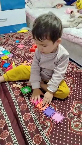 A young child sits cross-legged on a patterned rug, playing with colorful foam puzzle pieces. The child wears a long-sleeved, striped shirt and yellow pants. They are focused on connecting the puzzle pieces in a line. Various other toys are scattered around the rug. A bed with a white sheet and a pink trim is partially visible behind the child. To the left of the child is a blue and white dresser. The rug has a dark red base color with a repeating floral and geometric pattern.