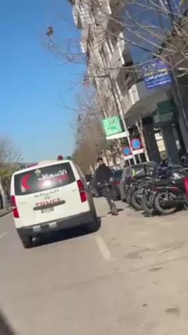A white police van, emblazoned with Arabic script and the word "POLICE" in English, sits parked on a street lined with parked motorcycles and vehicles. A man on an electric scooter passes in front of the van. A tall building is visible in the background, and street signs dot the scene under a clear, blue sky. The camera appears to be handheld and inside a vehicle.