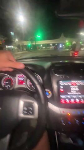 The video starts inside a car at night. The car appears to be a Dodge, based on the badge visible on the steering wheel. The driver's hand is visible on the steering wheel, waiting at a green light. The dashboard and infotainment system are visible, and a motorcycle is waiting on the right. The video abruptly transitions to a selfie view of the driver, a man in a blue sports jersey, briefly making a funny face.
