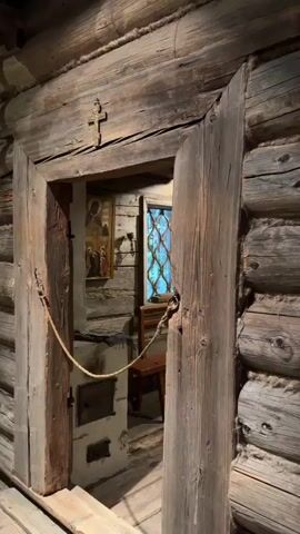 The video shows a view looking into a small, rustic room constructed of rough-hewn logs. A thick rope acts as a barrier, stretched across the doorway. The room appears to be a religious space. Above the doorway, a small cross is carved into the wooden beam. Inside, the walls are lined with numerous religious icons displayed on shelves. A wooden bench is placed against one wall beneath a window with a lattice pattern. To the left, a stove or oven is built into the wall, with a small metal door. The floor is made of wooden planks. The lighting is dim, suggesting an interior space. The overall impression is of a preserved historical or traditional dwelling or chapel.