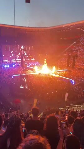 The video shows a large concert venue packed with an audience. The stage is illuminated with bright lights and pyrotechnics, including flames erupting from the stage. A large, golden, metallic statue of a person is prominently displayed on the stage, appearing to be suspended in the air. The audience members are holding up their phones, likely recording the performance. The overall atmosphere is energetic and visually dramatic.