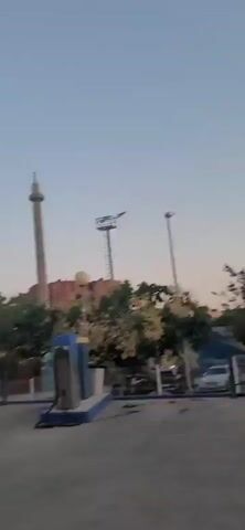 The video begins with a wide shot of an outdoor area with a tall tower in the background. The scene then transitions to a covered area, possibly a car wash or an outdoor cleaning station. A person wearing a black t-shirt, black shorts, a black baseball cap, and black shoes is seen using a pressure washer on a patterned rug lying on the concrete floor. The rug has a geometric border and a circular design in the center. Soap suds are visible on the floor around the rug. The person is holding the pressure washer wand and directing the stream of water onto the rug. Another person, wearing a green t-shirt, black shorts, glasses, and tan shoes, is also seen using a pressure washer on the same rug. The rug is being cleaned from multiple angles. The environment appears to be an open-air facility with a roof supported by pillars.