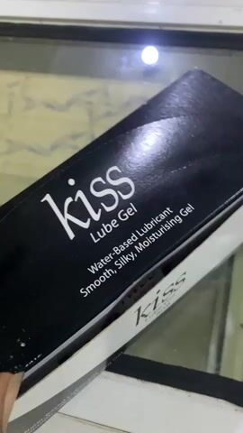 The video shows a person's hand opening a box of individually packaged Kiss Lube Gel sachets. The box is black with white lettering that says "Kiss Lube Gel Water-Based Lubricant Smooth, Silky, Moisturising Gel". Inside the box, there are rows of small, white and red foil packets, each containing 5ml of lube gel. The packets also have the Kiss logo and images of faces on them. The environment appears to be indoors, possibly on a reflective surface like glass or polished wood. The lighting is somewhat dim with a bright light source visible in the background.