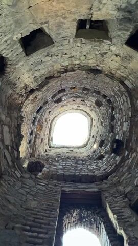 The video shows a view looking straight up inside a circular stone tower. The stone walls are rough and appear ancient, with many openings or windows at different levels. The structure appears to be spiraling upwards, creating a dizzying perspective. The top of the tower is open, revealing a bright sky. There is no evidence of people or activity within the tower.
