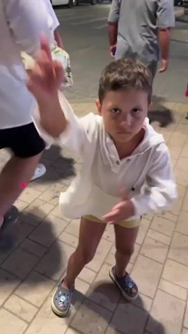A young child in a white shirt and yellow shorts dances on a sidewalk paved with beige interlocking bricks. The child's left arm is raised and bent at the elbow, hand outstretched, while their right arm is bent at the elbow and extended downwards. The child's legs are bent at the knees, with their left leg forward and their right leg behind. The child is wearing gray crocs with blue accents. In the background, several adults are walking on the sidewalk. Cars are visible on the street to the left of the frame, and a building with lights is visible in the distance. The lighting suggests it is evening or dusk.