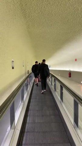 The video shows the back view of three men walking on a moving walkway in a tunnel. The walls of the tunnel are light yellow and textured, while the ceiling is a similar color and texture. There are small square pictures or signs on the walls, evenly spaced. The moving walkway has a grey, ribbed surface with glass barriers on either side, framed by silver railings. A fire extinguisher is visible on one side of the tunnel. The men are dressed casually. The first two are wearing black shorts or pants and jackets. The third man, closer to the camera, has a backpack and is wearing pants and sneakers with red and white accents. The tunnel appears well-lit. The men are walking toward the end of the tunnel, which seems to open into a brighter area.