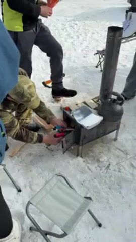Here's a detailed description of the video:

The video shows a person outdoors, in a snowy environment, attempting to light a small wood stove. The person, wearing camouflage clothing, is kneeling in front of the stove and using two handheld torches, one blue and one red, to ignite small pieces of wood inside.  

Around the stove, there are scattered pieces of wood, an axe lying on the snow, and several people standing or walking nearby. A metal kettle sits on top of the stove, and a foil container holding some kind of food sits on top of it as well. A portable folding stool is also visible near the stove.

The camera focuses closely on the person's hands and the stove as they work to light the fire. The background shows other people milling around, all bundled up for the cold weather, indicating an outdoor gathering in a winter setting.