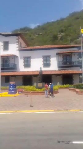 A brief video shows a street scene with a white building and a hill in the background. There is a motorcyclist with two passengers in the foreground, a person sweeping in the middle ground, and some greenery and a metal structure in the distance. The video seems to be taken from a moving vehicle.