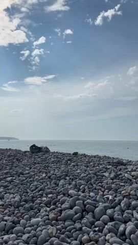 The video shows a rocky beach under a partly cloudy sky. The foreground is filled with smooth, gray pebbles. In the middle ground, the calm sea stretches to the horizon, meeting the sky. In the far distance, a faint coastline is visible. A few small figures can be seen in the water, possibly swimming or wading. The overall scene is tranquil and depicts a typical coastal landscape.