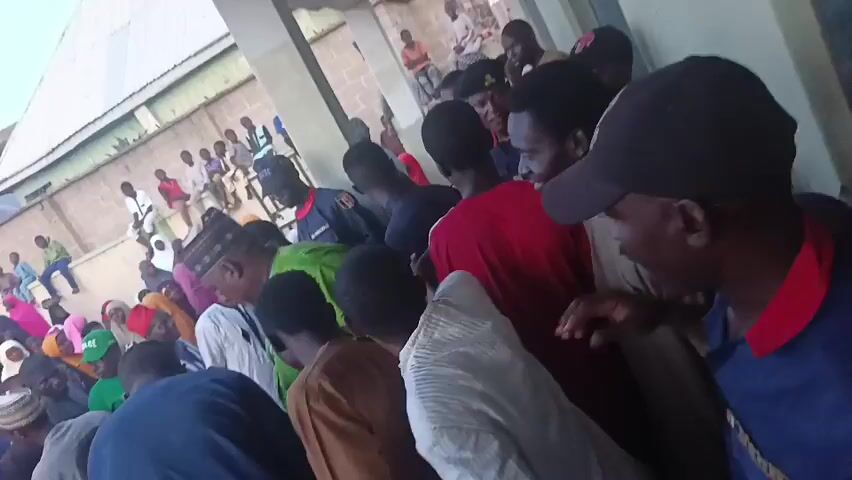 A large crowd of people, mostly men, are gathered in an outdoor space. Some people are standing close together in the foreground, while others are sitting on a wall or other elevated surfaces in the background. Most of the people are wearing casual clothing, and some of the men are wearing traditional hats or head coverings. The environment appears to be a residential area, with buildings and walls visible in the background. The camera is moving through the crowd, providing a close-up view of the people and the surrounding area. The overall atmosphere is one of gathering and community.