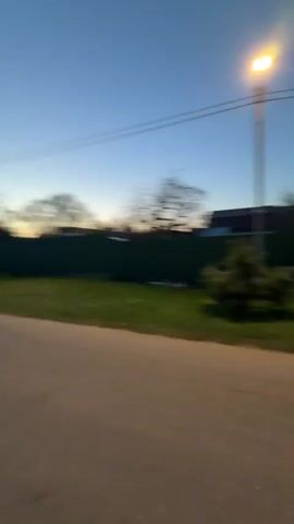 A young woman wearing a white long-sleeved shirt is the central figure in the video. She is standing on a paved road, facing the camera directly. She briefly makes a kissing face and then smiles.  Behind her, the setting suggests a rural or suburban area with houses, trees, and utility poles visible against a blue sky that appears to be either dusk or early morning. The road stretches into the distance, with vegetation bordering its sides. The camera seems to be hand-held, resulting in some movement and slight blurring.