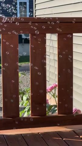 The video shows a wooden deck railing and part of a house. Bubbles are floating through the air, mostly in the upper left quadrant of the screen. Behind the railing, a pink rose is visible, along with green foliage. The siding of the house is light gray.