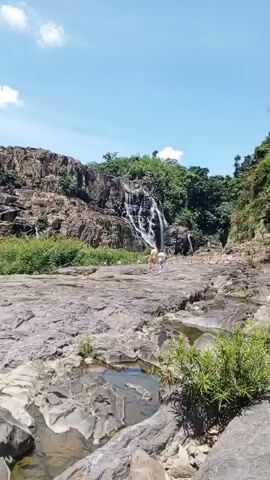 The video captures a wide shot of a cascading waterfall on a sunny day. The waterfall flows down a rocky cliff face, with lush green vegetation bordering the water and the cliff. In the foreground, there are large, flat rocks with smaller pools of water. Two figures, possibly tourists, are visible near the base of the waterfall, one wearing a yellow shirt. The sky above is blue with scattered white clouds. The overall scene depicts a natural, scenic environment.