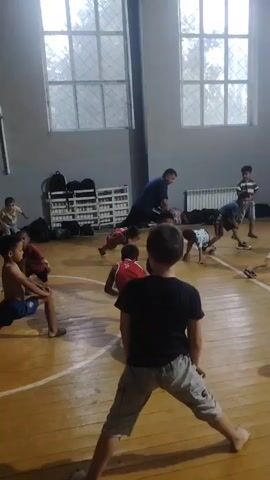 The video shows a group of young boys and a man in a gymnasium. The boys are dressed in athletic wear, including shorts and t-shirts, and one boy is wearing a red singlet with white trim. They are all engaged in a stretching exercise, with their bodies in various lunging and bending positions. The man, also dressed in athletic attire, appears to be instructing the boys, standing and observing their movements. The gymnasium floor is made of wood, and there are large windows with intricate grates on the far wall. Racks of equipment are visible against the wall, and a radiator is also present. The overall atmosphere suggests a sports training session or physical education class.