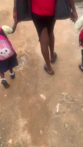 A person wearing a red shirt and black skirt walks along a dirt path, holding hands with two children. The child on the left wears a yellow shirt, a dark blue skirt, and a pink backpack with a white bunny design. The child on the right is partially visible and also appears to be wearing a yellow shirt and dark uniform. The person is carrying two dark-colored bags, one in each hand. The path is uneven and dusty, with some gravel and small puddles visible. A black car is parked in the background.