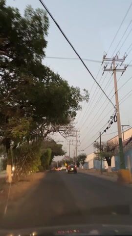 The view from the dashboard of a vehicle driving down a paved road lined with trees, bushes, and power lines. Several cars are visible ahead, including one dark-colored sedan. A large building with a blue wall stands to the right of the road. The sky is overcast.
