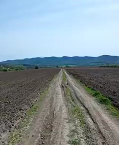 The video begins with a landscape shot of a dirt road cutting through a large plowed field. In the distance, green hills are visible under a blue sky with scattered clouds. The camera then shifts to a close-up of the handlebars of a bicycle, showing the blue frame. It then returns to the landscape view, moving to the right, maintaining the focus on the plowed field, dirt road, and the distant hills under the same sky conditions.