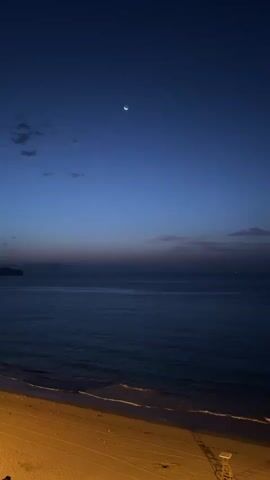 The video shows a twilight view of a beach and the ocean under a dark blue sky with a crescent moon. The beach is sandy and appears to be lit by artificial lights. The ocean is dark blue with gentle waves lapping at the shore. The sky transitions from a deep blue at the top to lighter shades of blue and purple near the horizon. There are a few wispy clouds visible in the sky. In the distance, there appears to be land or a coastline. There are no people or animals visible in the frame. There is a lifeguard tower on the right side of the beach. The video is still and does not show any movement.