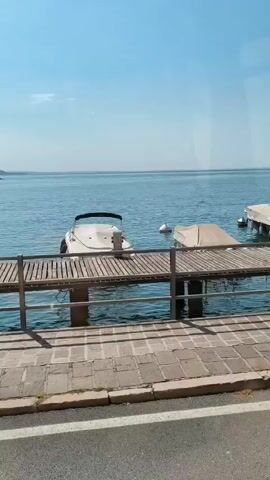 The video opens with a wide shot of a calm blue lake under a clear sky. Three boats are docked at a wooden pier. The boat on the left is white with a black canopy. The middle boat is covered with a beige tarp. The boat on the right is also covered. A person is visible on the rightmost dock, standing and facing the boat. The camera then pans to the left, showing a blue van driving past. The driver, wearing a dark shirt and a hat, is visible inside the van. The video then returns to the lake scene, with the person on the right dock still standing and appearing to adjust the cover on the boat. The camera continues to pan left, briefly showing the side of the van. The video ends with a shot of the lake and the docked boats.