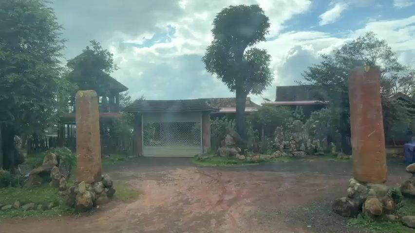 The video shows a view from a moving vehicle traveling along a dirt road. On the left side of the road, there are two tall, weathered stone pillars standing upright, surrounded by piles of smaller rocks and grass. Beyond these pillars, a gate is visible with a rolling shutter, and behind it, a building with a reddish roof. A large tree with green foliage stands to the left of the gate.

Further along the road, on the right side, a unique structure with a conical roof and stone-like textured walls resembles a small tower or a whimsical building. Palm trees are scattered throughout the scene, and the ground is a mixture of dirt and patches of grass. Various rock formations and decorative landscaping are present along the roadside.

In the background, glimpses of other buildings, including a large industrial-looking structure with a tall billboard on top, are visible. People are seen occasionally, including a person on a motorcycle, and small groups of individuals standing around. The sky is cloudy, suggesting an overcast day. Red boats are also visible in the distance. The overall impression is of a rural or semi-rural area with elements of unique landscaping and architecture.