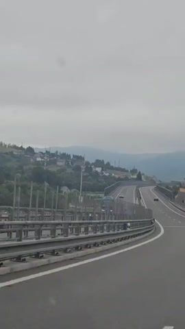 This video shows a POV shot from a vehicle traveling on an elevated highway. The highway has multiple lanes and is divided by white dashed lines. There are concrete barriers on either side of the road, with metal railings in front of them. A blue bus is visible on the highway in the distance, as well as several cars. The landscape to the left of the highway consists of rolling hills covered in trees and dotted with houses and buildings, suggesting a semi-rural or suburban environment. Mountains are visible in the background, shrouded in a light haze under a cloudy sky. The sky is overcast, indicating a gray or possibly rainy day. The highway appears to be ascending a gentle slope.