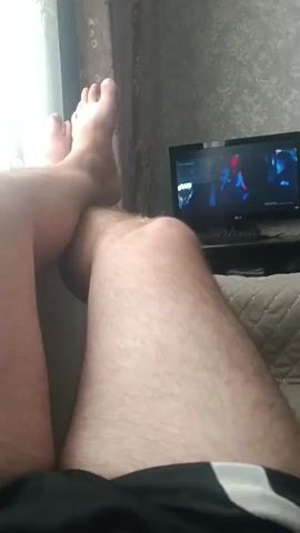 The video shows a first-person perspective of a person's legs and feet. The person is lying down with their legs crossed, and one foot is resting on the other leg. The person is wearing black shorts. In the background, a television screen is visible, displaying an image that appears to be from a video game. The room is dimly lit, with light coming from a window on the left side of the frame. The overall scene suggests a person relaxing and watching television.