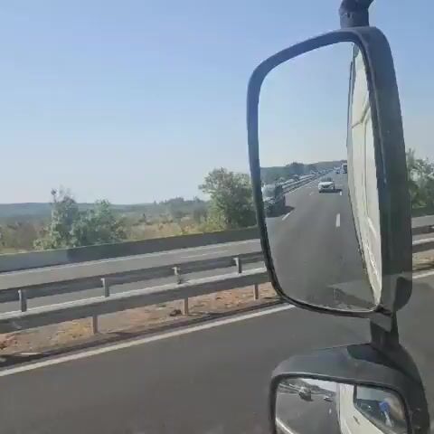The video captures a view from the inside of a vehicle driving on a highway. The primary focus is on the road ahead and the surrounding environment. The highway appears to have multiple lanes, with traffic visible in the distance. There are other vehicles, including cars and trucks, on the road. A police car with flashing blue lights is also present, indicating a potential traffic stop or incident. Trees and vegetation line the side of the highway, and the sky is clear and blue, suggesting a sunny day. The video also includes shots from a side mirror, showing a clearer view of the traffic behind. The overall scene depicts a normal highway environment with a police presence.