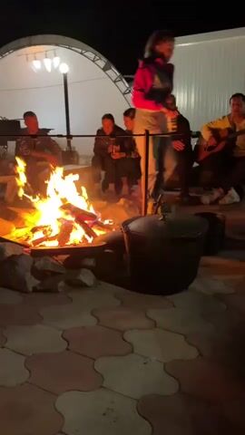 A group of six people are gathered around a bonfire at night. The fire is in the center of the frame, with flames and smoke rising. To the left of the fire, three people are sitting. To the right of the fire, three people are sitting or standing. One person in the foreground, on the right, is standing and walking. Two large pots are placed near the bonfire. The ground is paved with interlocking tiles. In the background, there is a white building and a white arched structure with lights. The overall atmosphere appears to be social and relaxed.