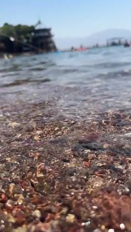 The video shows a close-up, low-angle view of a rocky shoreline with clear water gently lapping over small pebbles. In the background, the water extends to a hazy horizon with several indistinct figures and structures visible, suggesting a beach or resort area. The sky is a clear, pale blue. The focus is on the pebbles and the shallow water, with the background blurred.