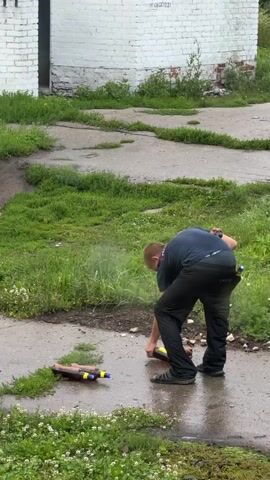 The video shows a young man in a dark blue t-shirt with the word "ПОЛИЦИЯ" (Police) on the back and dark pants. He is standing on a wet paved surface, with overgrown grass on either side. In front of him, there are three objects lying on the ground that appear to be bottles with yellow and blue accents. The man is holding two brown bottles, one in each hand. He appears to be spraying something from the bottle in his right hand, creating a mist. He is looking down at the bottles on the ground. The background shows a white brick building with some graffiti and overgrown vegetation. The overall scene suggests an outdoor, somewhat neglected urban or semi-urban environment.