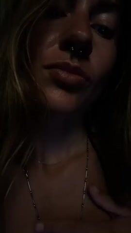 A close-up video shows a person, likely female, from the chin to the forehead, with long, light-colored hair partially visible on the sides. She has a septum piercing and is wearing two necklaces, one thin chain and one thicker chain with a hook-shaped clasp. A simple silver ring is on her left ring finger.

The video begins with the person looking towards the camera. She then slowly brings her left hand up to her throat, then her chin, and finally her fingers reach her lips, which are slightly parted in a pouting or kissing gesture. The lighting is low, illuminating her face from above and casting shadows, suggesting a dark indoor setting or nighttime. The background is completely dark. The overall mood appears playful or suggestive. There are no visible location cues or text.