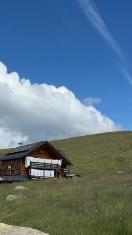 A rustic wooden cabin sits nestled on a grassy hillside under a bright blue sky with fluffy white clouds. Laundry is hung to dry on the balcony of the cabin. The cabin has a dark roof and a chimney. A cross is visible on the hilltop in the distance. The foreground is filled with tall green grass. The video is a static shot of the scene.