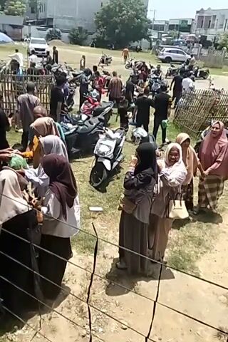 The video shows a gathering of people outdoors, possibly at an event or a public space. In the foreground, several people are visible, some wearing Islamic attire like hijabs and long dresses. One person in a black shirt is walking up stairs, carrying two blue objects that appear to be bags or containers. Other individuals are standing around, some interacting with each other. In the background, there are more people, vehicles including motorcycles and a white car, tents, and some structures that resemble buildings. The scene suggests a bustling, community-oriented environment, possibly a religious gathering or a social event.