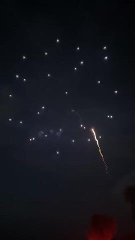 The video shows a fireworks display at night. Several fireworks are visible in the dark sky, some are exploding in vibrant colors like red and yellow, while others are still ascending or fading. The fireworks are the primary focus, with the sky serving as the background. There are also some distant lights or stars visible. At the bottom right of the frame, a dark, possibly silhouetted object or structure is partially visible, along with a faint red glow. The overall environment is dark, indicative of nighttime. No people are clearly visible in the frames.