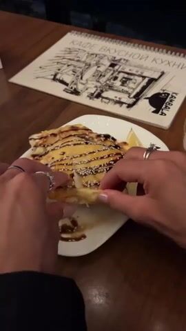 A close-up shot from an overhead perspective shows a person's hands tearing into a dish of food. The dish is on a white plate and appears to be some kind of savory pastry or bread filled with a creamy, possibly cheesy, filling. The filling is visible as it stretches and pulls away from the pastry. The pastry is golden brown and topped with sesame seeds and a dark sauce drizzle. A slice of lime is visible on the side of the plate. In the background, a menu or booklet with a drawing on it is placed on a wooden table. The lighting is dim, suggesting an indoor setting, possibly a restaurant. The person's hands are adorned with rings, and their fingernails are painted. The action of tearing the food is the main focus of the video.