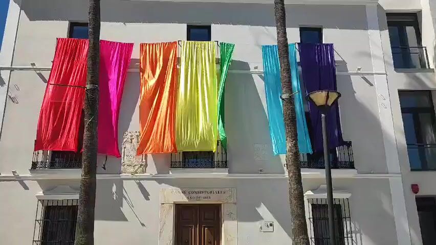 The video shows a medium shot of a white building with several large, colorful fabric banners hanging from balconies on the second floor. The banners are arranged in a rainbow pattern, with red, pink, orange, yellow, green, light blue, and purple fabrics visible. Two palm trees frame the scene in the foreground. The building has several windows with dark shutters and a central entrance with a wooden door. There is a lamppost to the right of the banners. The sky is blue and the sunlight casts shadows on the building.