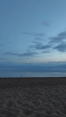 The video shows a static shot of a beach at dusk. The sky is a deep blue with some scattered clouds. A sliver of the moon is visible in the upper center of the frame. The ocean is calm and stretches to the horizon. The foreground is filled with dark, rippled sand, typical of a beach. A few small red buoys are dotted along the waterline, suggesting a designated swimming area or the presence of some form of aquatic activity. There are no people or animals visible in the frame. The overall mood is serene and tranquil.