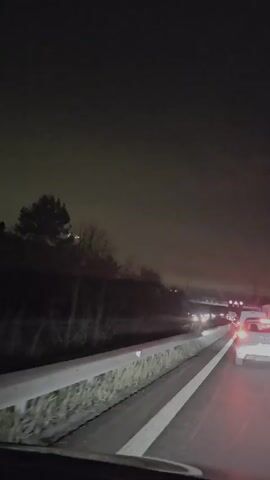 The video shows a car driving on a highway at night. Other cars are visible ahead in both lanes, some with their brake lights on. A concrete barrier separates the road from a dark wooded area. The sky is dark with a slight glow from the city lights.  There's music playing. The camera appears to be inside a vehicle, possibly on the dashboard.
