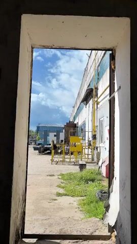 The video shows a view through an open doorway onto an outdoor industrial or warehouse area. The setting appears to be a sunny day with some clouds, as indicated by the bright sky and shadows. The main focus is the exterior of a long, grey building with blue-tinted windows and yellow pipes running along its side. In front of this building, there is a yellow fence, some vehicles, and piles of debris. A blue building can be seen in the background. There are no people visible in the frame. The overall impression is one of a functional, possibly somewhat neglected, industrial environment. There is some visible text on a white sign attached to the yellow fence, but it is too small to be legible in the video. The time of day appears to be daytime.