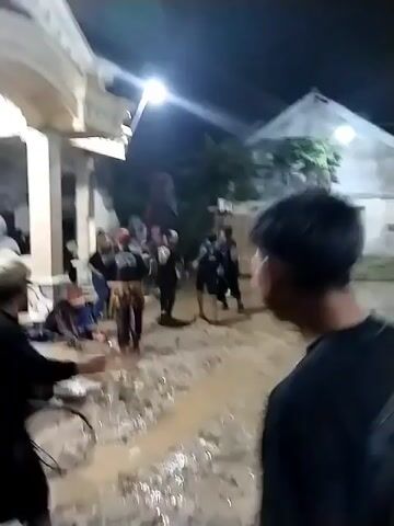 At night, an outdoor area in Jenggrik, Indonesia, is illuminated by a bright overhead light, revealing a muddy ground with puddles. A group of people, primarily young adults, are gathered. Many wear black t-shirts with yellow or gold emblems on the back. A light-colored house with columns is visible on the left, while wooden structures line the background on the right. In the distance, individuals are actively moving, and what appears to be a large, dark figure, possibly a performance prop, is being maneuvered. The sound of music and drumming suggests a lively event or performance is taking place, with participants moving through the wet, muddy conditions.