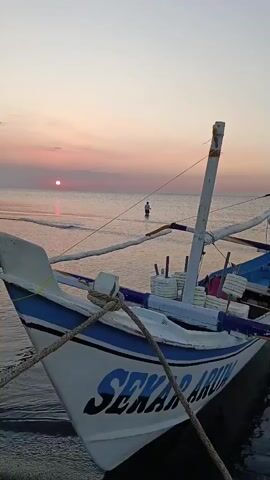 The video shows a close-up of a white fishing boat with blue trim and the name "SEKAR ARUM" written on its side, anchored near the shore. The boat is positioned at an angle, facing away from the viewer and slightly to the right. The water is calm with gentle waves lapping at the shore. In the background, the sun is setting over the ocean, casting a warm glow on the sky, which transitions from pink to orange. A lone figure, appearing to be a person, stands in the shallow water further out from the shore, facing the sunset. The overall scene depicts a peaceful coastal setting at dusk.