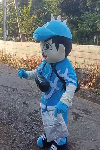 A person in a blue mascot costume, resembling a cartoon character, stands outdoors on a gravel and dirt path. The mascot has a large head with black hair, large white eyes with black pupils, and a wide smile. It wears a blue jumpsuit with white accents and blue gloves. A black cross-body bag is worn over the costume. The mascot holds a crinkled silver object in its right hand and appears to be interacting with a young person partially visible on the right side of the frame. The background includes green foliage and a low brick wall. The video is horizontally oriented.